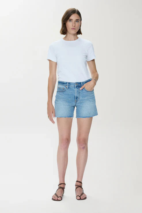 A woman with short brown hair stands against a plain light background, wearing a white short-sleeve tee and Zertz KENNEDY RELAXED MID RISE CUT OFF SHORT in sun-washed blue denim, paired with black strappy sandals and one hand in her pocket.