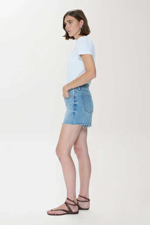 A person with short brown hair stands in profile against a white background, hands in pockets, wearing a white T-shirt, Zertz KENNEDY RELAXED MID RISE CUT OFF SHORT in sun-washed blue denim, and brown strappy sandals.