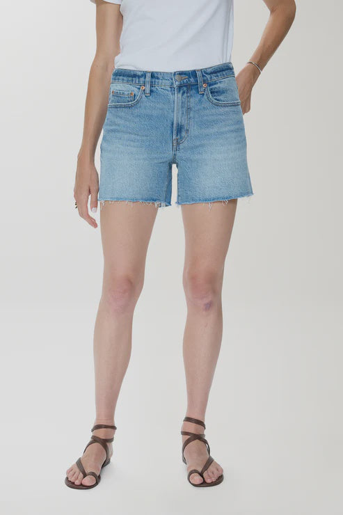 A person stands against a plain light background wearing a white t-shirt, brown strappy sandals, and Zertz KENNEDY RELAXED MID RISE CUT OFF SHORTS in sun-washed blue comfort stretch denim with frayed hems.