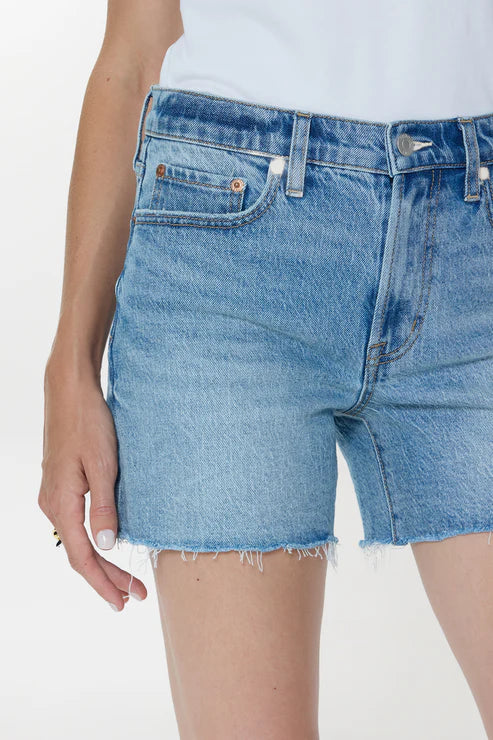 A person wears Zertz KENNEDY RELAXED MID RISE CUT OFF SHORTS in sun-washed blue comfort stretch denim and a white top, with their left hand resting by their side.