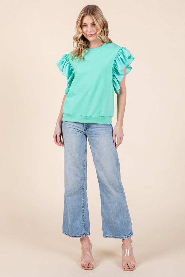 A woman stands smiling against a beige background, wearing the mint green Rufflina Top by Loca on Main with short sleeves, paired with light blue wide-leg jeans and clear heeled sandals.