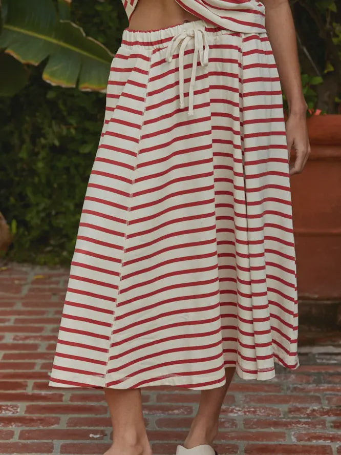 A person wearing the Zertz SASHA STRIPE KNIT SKIRT, featuring red stripes and a drawstring waist, stands on a brick pathway surrounded by greenery.