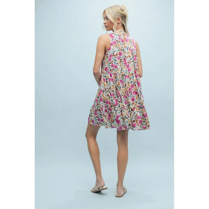 A woman with blonde hair in a loose updo models the SLVLESS PATTERN DRESS by Zertz, a floral satin mini dress with pink, red, and green accents. She stands turned away in white sandals against a light blue background.