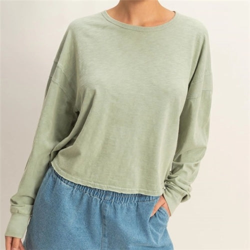 A person in a Loca on Main Laeme Open Back Top (light green, long sleeve) and high-waisted blue elastic denim pants stands with one hand in their pocket against a plain light background.