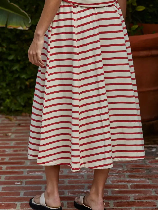 A person stands on a brick patio wearing the cream SASHA STRIPE KNIT SKIRT by Zertz with red horizontal stripes and white slide sandals, surrounded by greenery and a large terracotta pot.