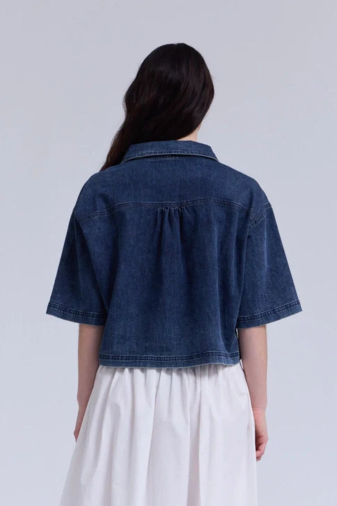A person with long dark hair, seen from behind, wears the Zertz DENIM CROP SHIRT over a white gathered skirt or dress, set against a plain light background.
