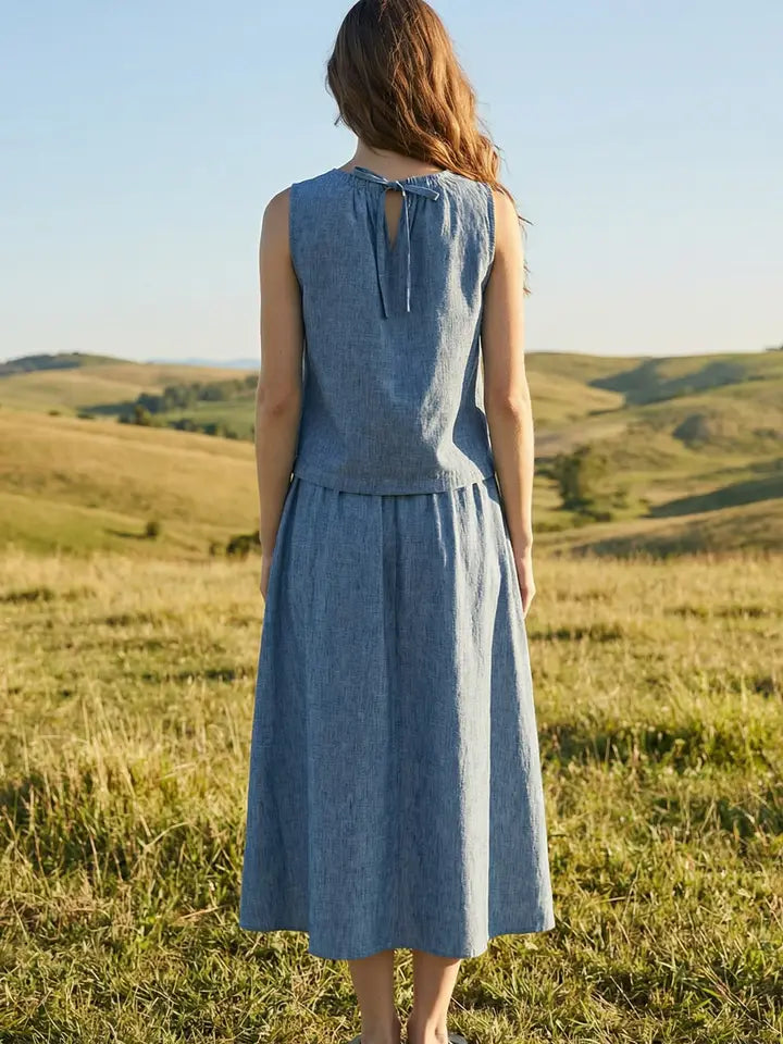 A woman with long wavy hair stands in a grassy field, wearing the Zertz SUNSET EMBROIDERY SET—a sleeveless, light blue dress featuring embroidered patches at the neck. Rolling hills and clear skies complete the serene scene.
