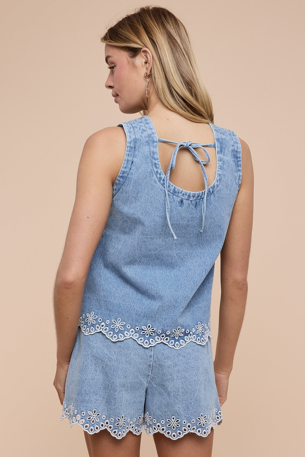 A woman with long blonde hair wears the Louisa Denim Set by Loca on Main—a light blue, sleeveless tank top and shorts with white floral embroidery—tying at the back as she faces away from the camera against a beige background.
