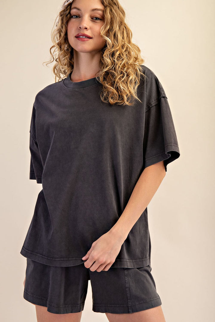 A person with long, wavy blonde hair models The Mellow Yellow Set by Loca on Main—featuring a mineral washed tee and relaxed fit dark gray shorts—while standing against a plain beige background.