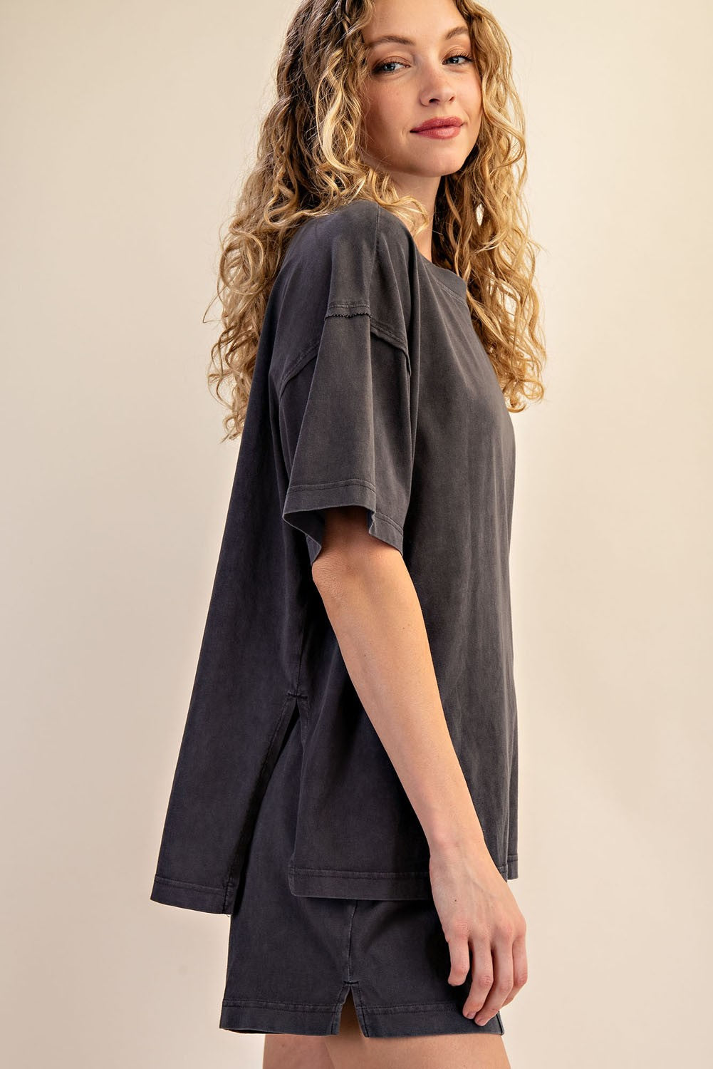 A woman with curly blonde hair wears The Mellow Yellow Set from Loca on Main, a relaxed fit shorts set and mineral washed tee in dark gray. She stands sideways against a beige background—ideal for comfortable everyday loungewear.