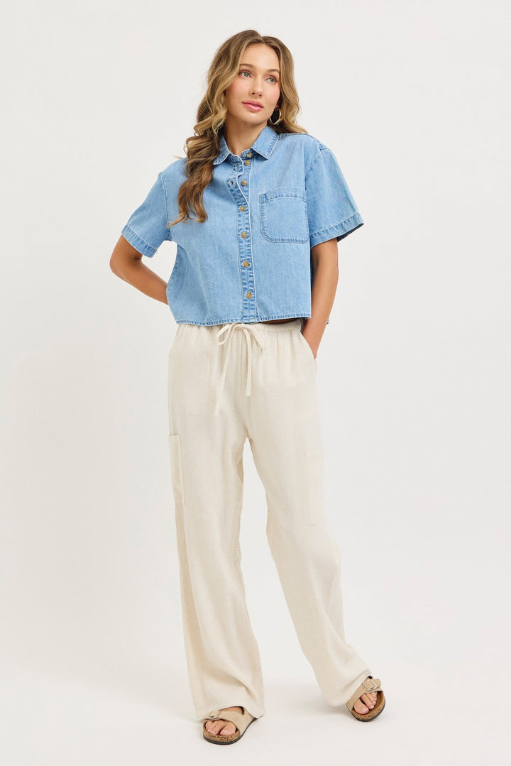 A woman with long, wavy hair poses relaxedly with one hand in her pocket, wearing The Jen Denim Top by Loca on Main against a plain background, paired with loose cream drawstring pants and sandals.