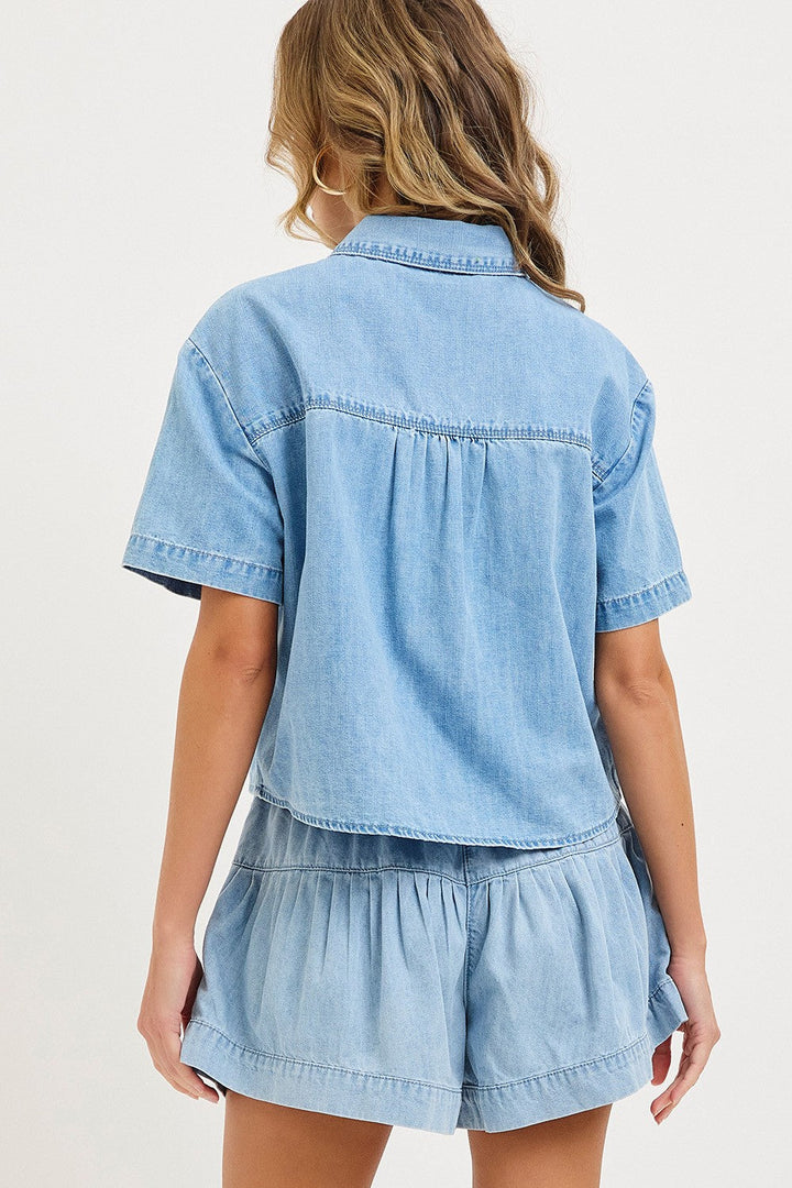 A person with wavy, light brown hair is shown from behind wearing The Jen Denim Top by Loca on Main—a light blue, washed shirt paired with matching high-waisted denim shorts. The background is plain and light-colored.