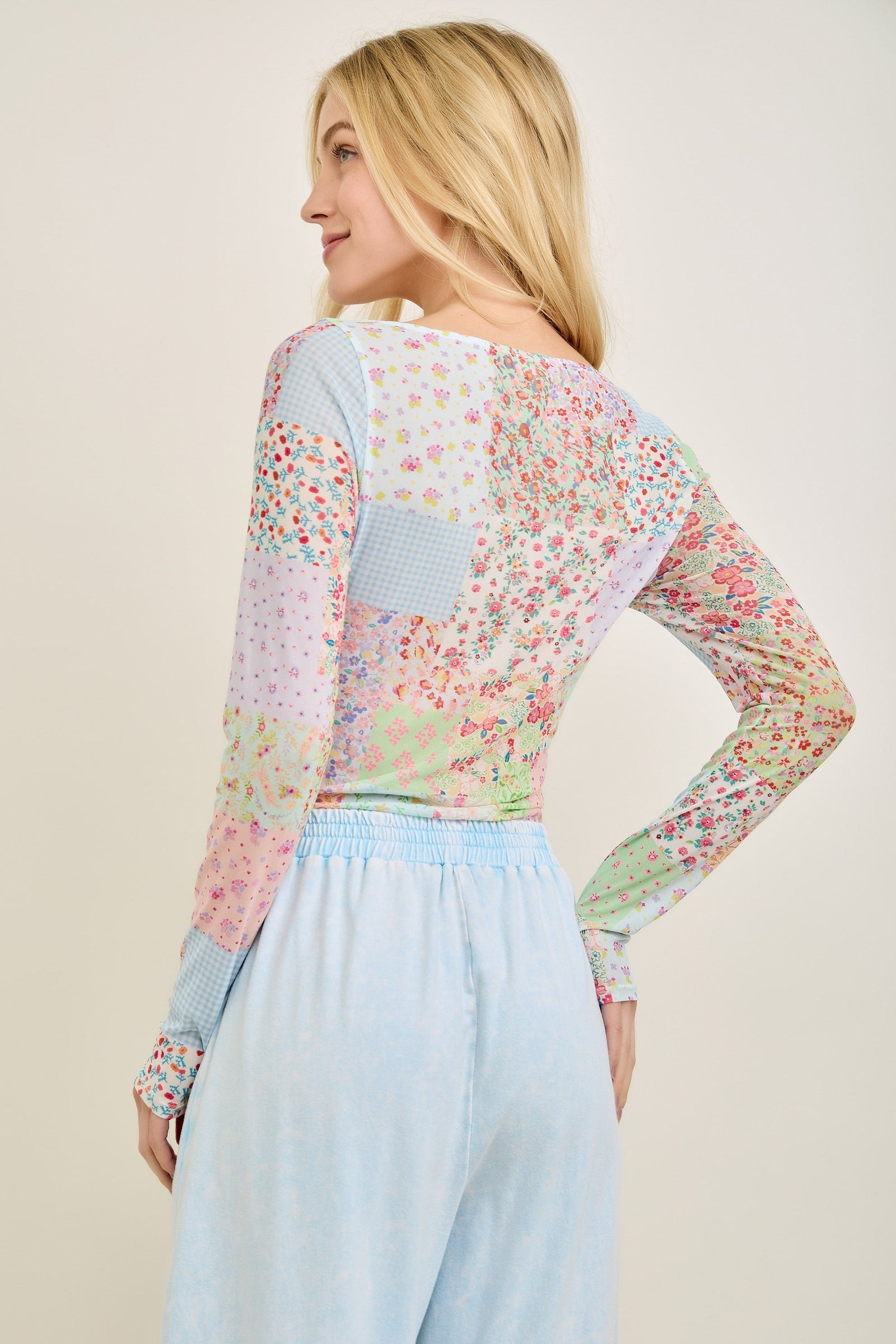 A woman with long blonde hair, seen from the back, smiles while wearing the Loca on Main Picnic Patch Mesh Top and light blue high-waisted pants against a plain light background.