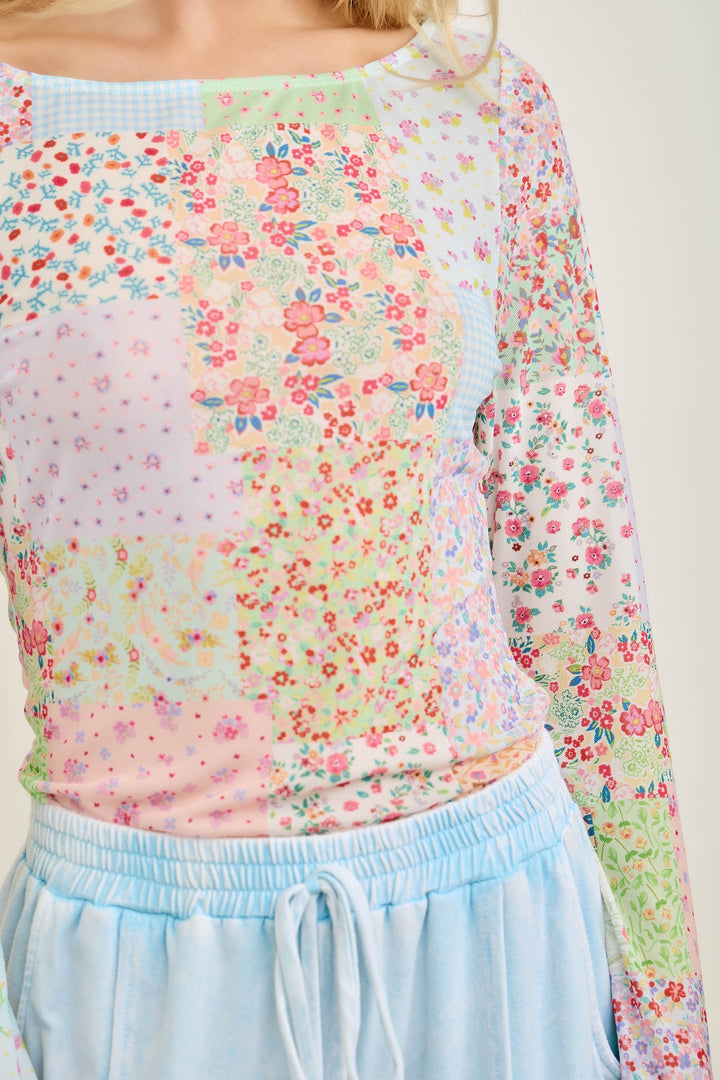 A person is wearing the Picnic Patch Mesh Top by Loca on Main, featuring a colorful floral patchwork pattern, paired with light blue drawstring pants. The image is cropped from shoulders to hips to showcase the outfit.