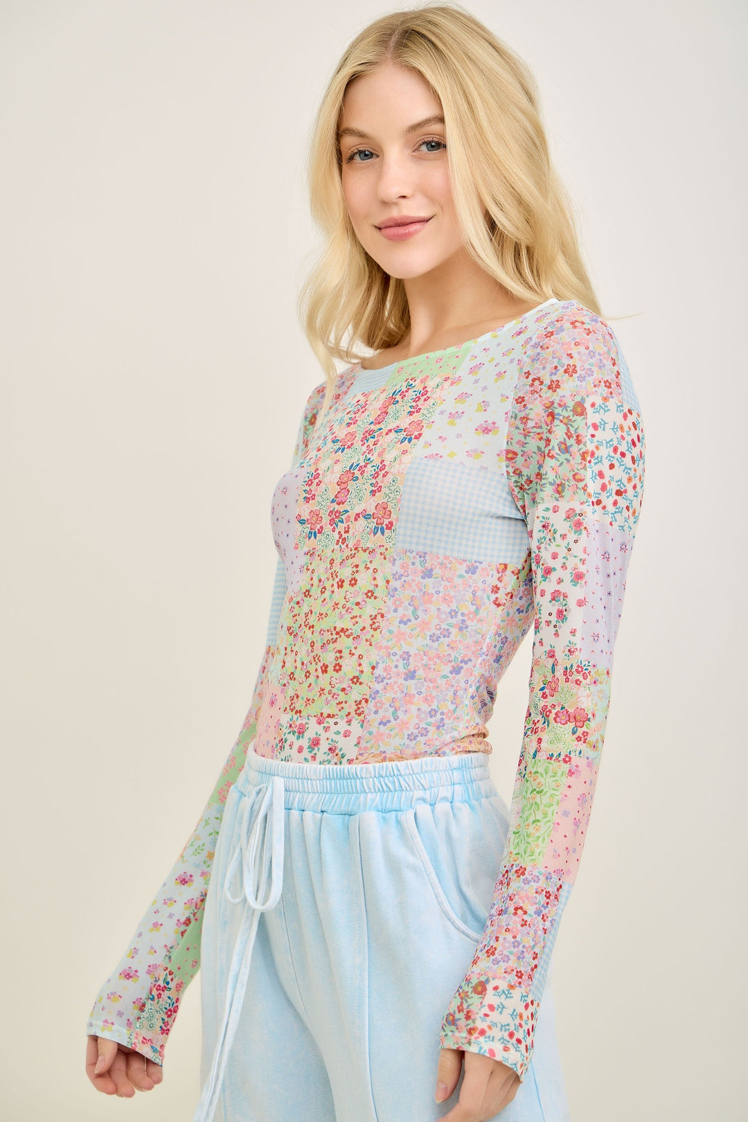 A woman with long blonde hair wears the Loca on Main Picnic Patch Mesh Top and light blue drawstring pants, standing against a plain light background and smiling slightly at the camera.