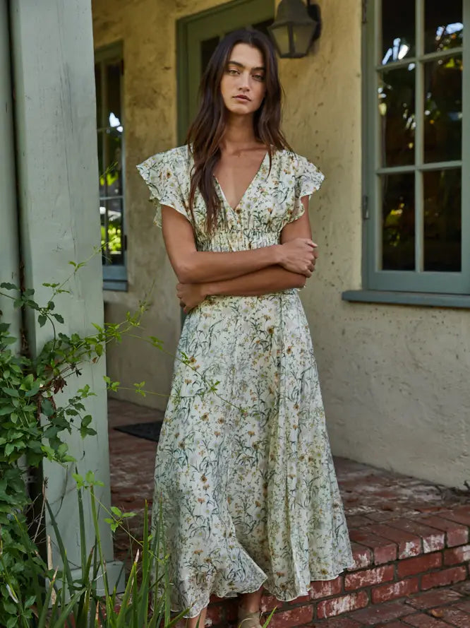A woman with long brown hair stands outside a house with green window frames, wearing the Zertz SEREIA FLORAL TOP—featuring short ruffled sleeves and a V-neckline—as she crosses her arms and looks at the camera.