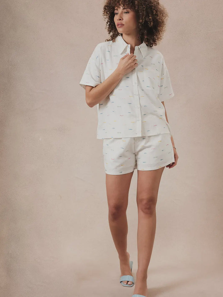 A person with curly hair models Zertz FISH PRINTED SHORTS and a light button-up shirt, standing against a neutral background. They hold the collar and wear open-toe slippers, effortlessly showcasing summer style.