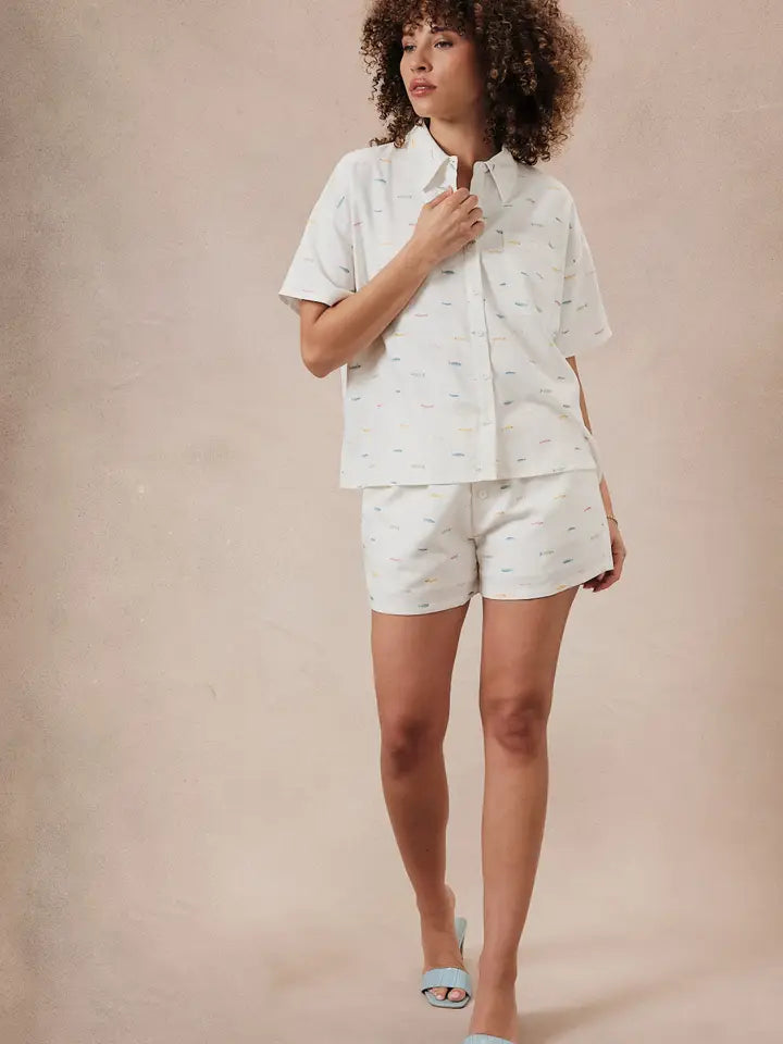 A woman with curly hair wears the Zertz PRINTED FISH SHIRT and matching shorts, featuring a subtle pattern. She stands against a neutral backdrop, adjusting her collar and evoking coastal vibes as she looks to the side.