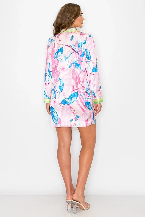 A woman with light brown hair stands facing away, wearing Zertz’s FLORAL PRINTED SHIRT DRESS featuring a pink, blue, and green tropical leaf pattern, styled with clear high-heeled sandals against a plain white background.