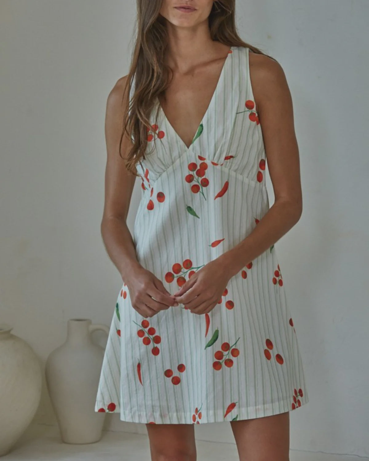 A woman wears the Zertz CAPRESE MINI DRESS—a sleeveless, V-neck mini dress with vertical stripes and a red cherry print—standing indoors with hands clasped, exuding effortless summer style. White ceramic vases are in the background.