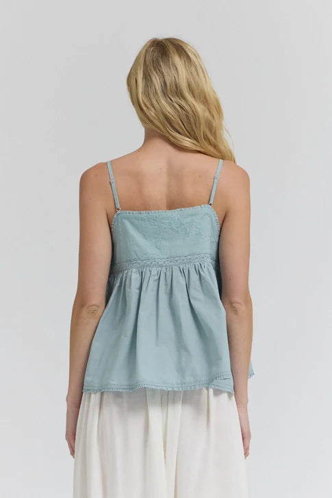 A woman with long blonde hair is seen from the back wearing the Zertz EMBROIDERED SLEEVELESS TOP—a light blue, thin-strapped top with a flared hem—paired with a white skirt against a plain light background.