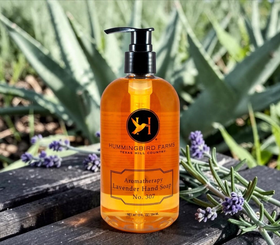 A bottle of Hummingbird Farms Lavender Aloe Hand Soap No. 307 rests on a rock outdoors, framed by lavender and aloe plants, highlighting its soothing, natural cleansing experience.