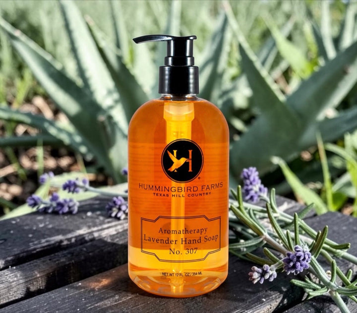 A bottle of Hummingbird Farms Lavender Aloe Hand Soap No. 307 rests on a rock outdoors, framed by lavender and aloe plants, highlighting its soothing, natural cleansing experience.