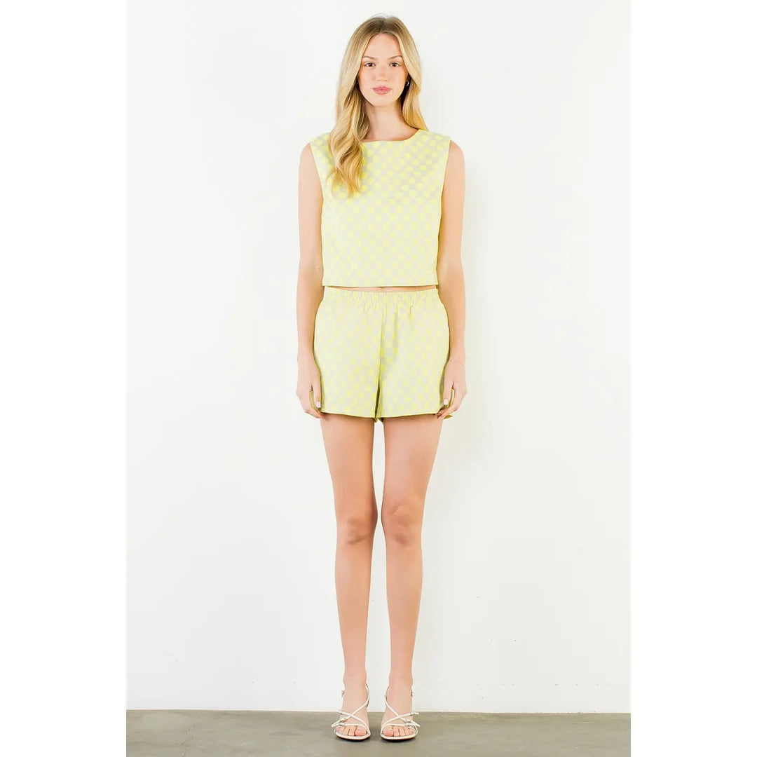 A woman with long blonde hair and a neutral expression stands against a white wall, wearing the Zertz POLKA DOT TOP and matching shorts for a comfortable fit, paired with white sandals.