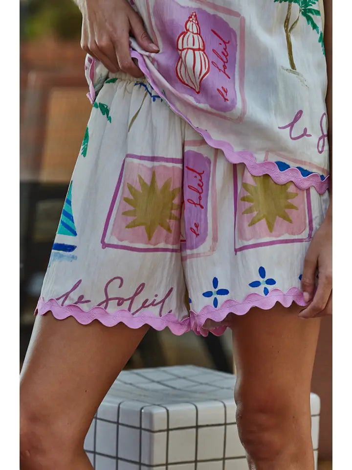 Someone stands near a tiled surface, wearing Zertz MONTMARTRE WOVEN SHORTS—white elastic waist shorts with pink trim and colorful prints like a starburst, seashells, and waves—with one hand resting on their hip.