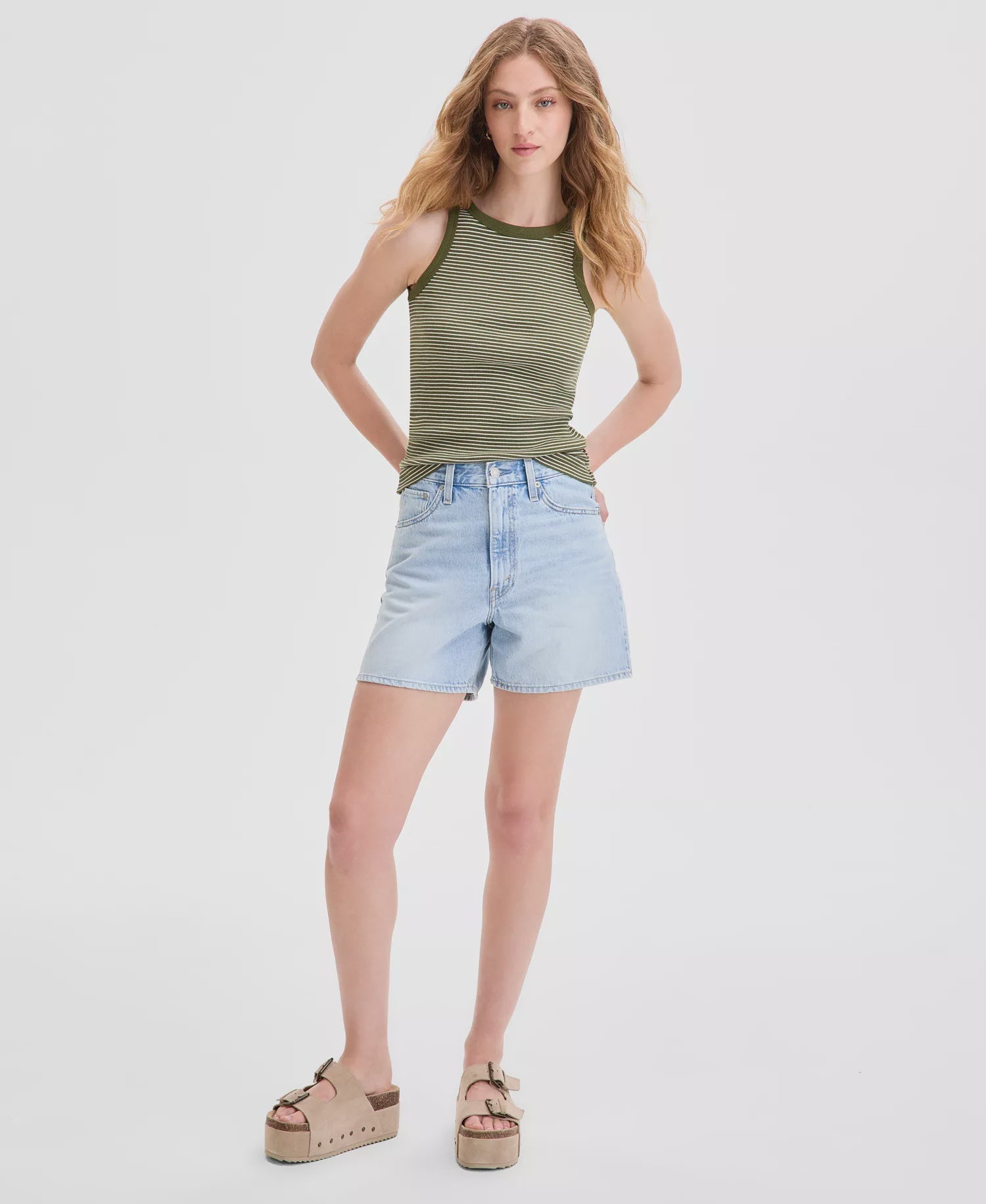 A woman with long, wavy hair stands against a white background, wearing Zertz CINCH MID-THIGH SHORT ABRAIDED ART high-rise baggy denim shorts with an adjustable waist, a green striped tank top, and beige platform sandals.