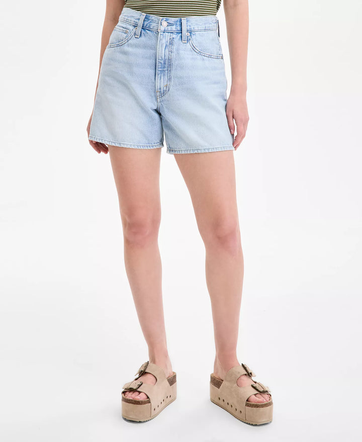 A person wearing Zertz CINCH MID-THIGH SHORT ABRAIDED ART in light blue denim with an adjustable waist, paired with a green striped top and tan buckle platform sandals, stands against a plain white background.
