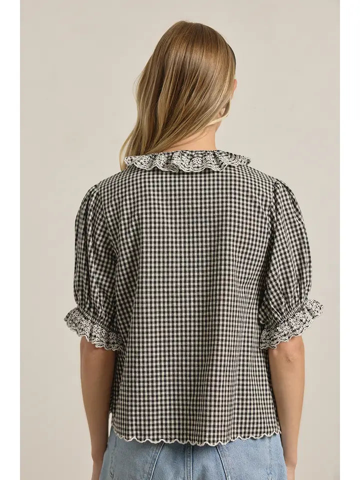 A woman with long blonde hair, facing away, wears the Zertz GINGHAM LACE RUFFLE BLOUSE—a vintage-inspired black and white gingham top with lace-trimmed collar and sleeves—paired effortlessly with light blue denim shorts.