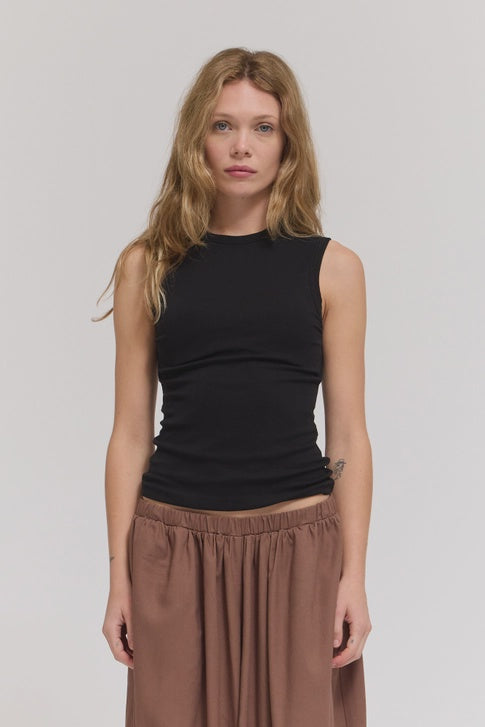 A woman with long, wavy blonde hair stands facing forward, wearing the Zertz SHEER TANK TOP in black and a loose brown skirt against a plain light background.