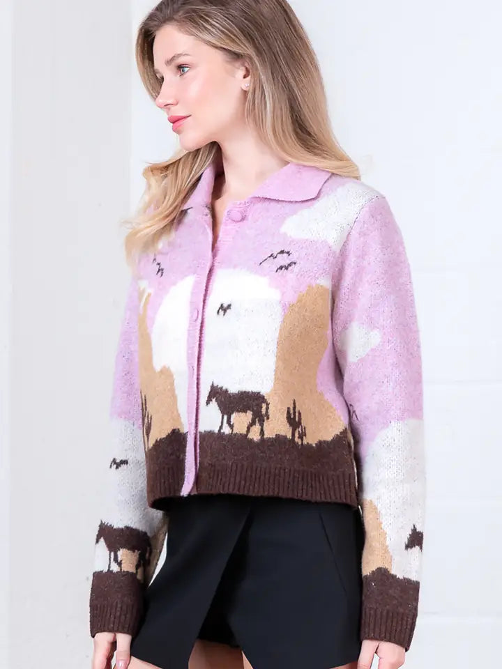 A woman with light brown hair stands sideways, looking left, wearing the Zertz JESSIE HORSE KNIT SWEATER in pink and beige with desert scenery—horses, cacti, and birds—and a black skirt.
