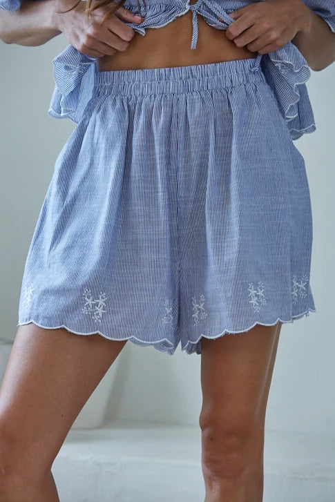 A person wears the Zertz CELESTINE SHORTS—loose, light blue and white striped shorts featuring a ruffled waistband, scalloped hems, breathable fabric, and delicate white floral embroidery along the edges.