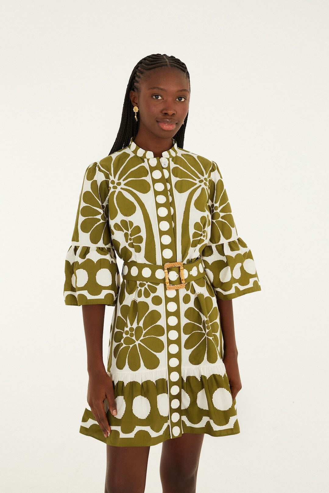 A woman with long braided hair and gold earrings stands against a plain background wearing the Zertz PALERMO GREEN DRESS, featuring bold florals, bell sleeves, a belted waist, and decorative buttons in organic cotton.