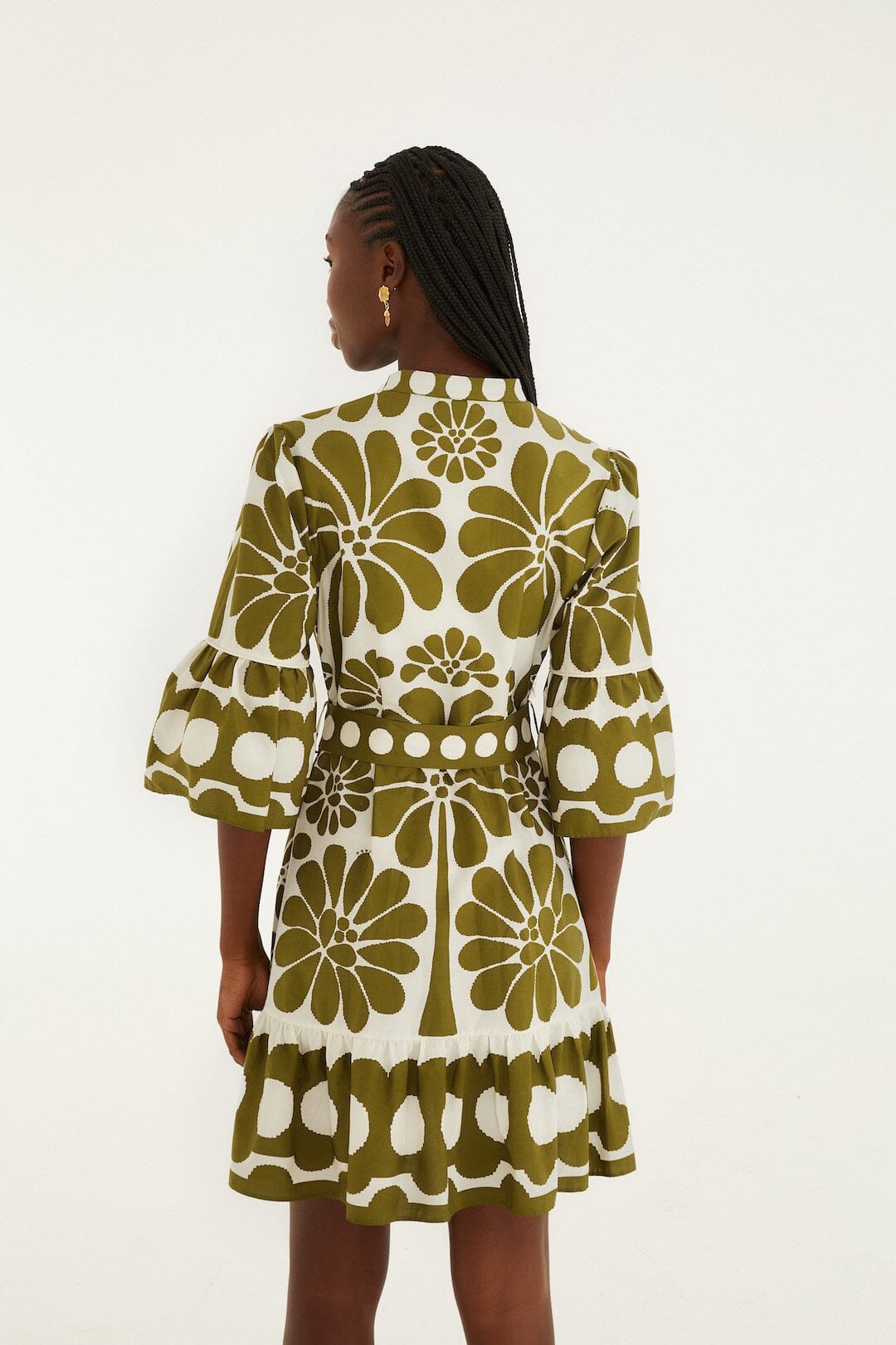 A woman with braided hair and gold earrings wears the Zertz PALERMO GREEN DRESS, featuring bold green floral and dot patterns, as she stands facing away against a plain white background.