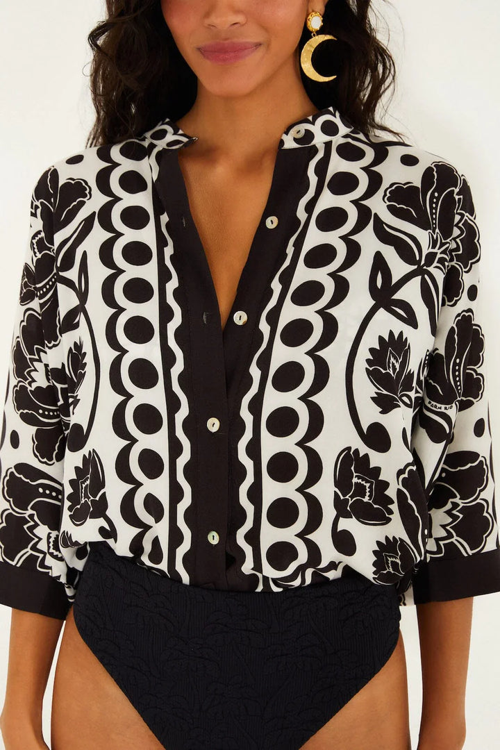 A woman wears a Zertz FLORAL ARABESQUE SHIRT with bold black and off-white florals. She pairs it with large gold earrings, one crescent moon-shaped, and black high-waisted bottoms. Only her upper body is visible.