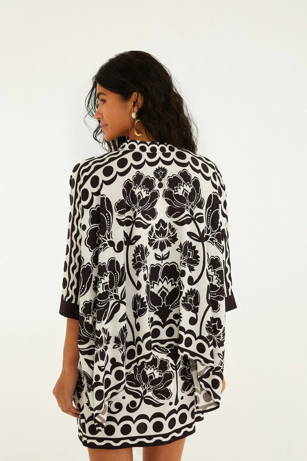 A woman with long, wavy dark hair stands facing away, wearing a Zertz FLORAL ARABESQUE SHIRT featuring black and white floral patterns and circular borders. She accessorizes with gold hoop earrings against a plain white background.