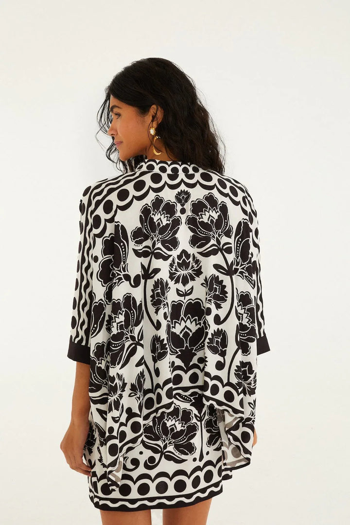 A woman with long, wavy dark hair stands facing away, wearing a Zertz FLORAL ARABESQUE SHIRT featuring black and white floral patterns and circular borders. She accessorizes with gold hoop earrings against a plain white background.