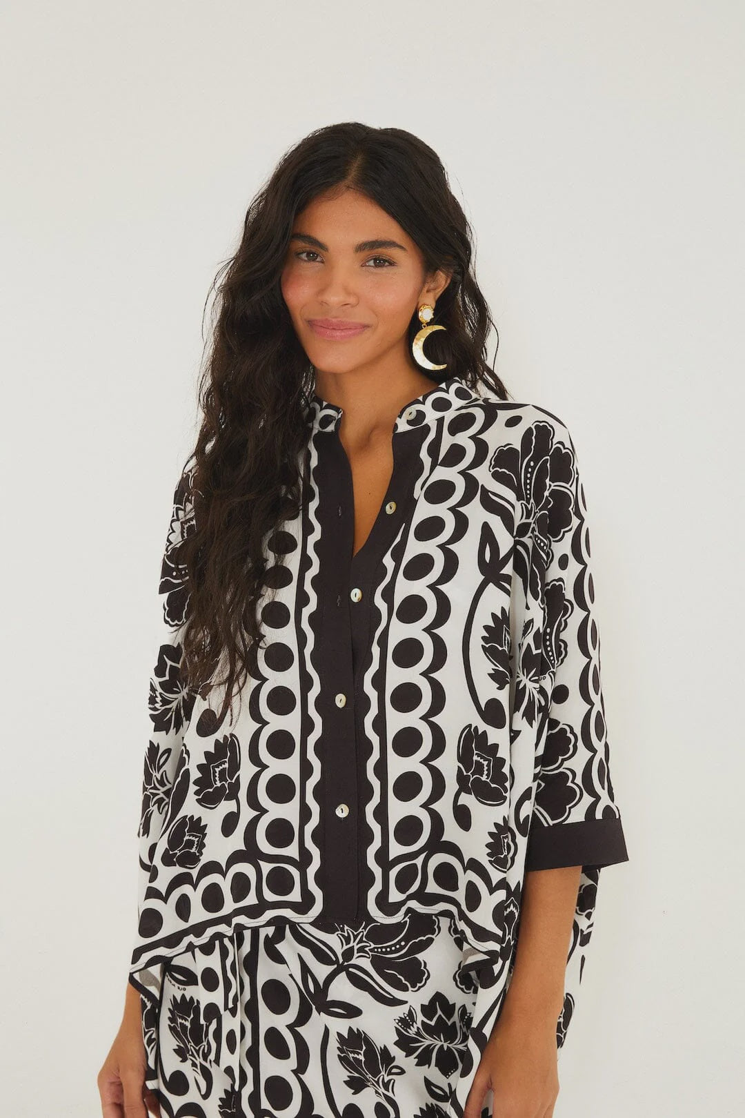 A woman with long, wavy dark hair wears a Zertz FLORAL ARABESQUE SHIRT in off-white with floral and geometric patterns. She stands against a white backdrop, smiling gently and showcasing large round earrings in a confident pose.