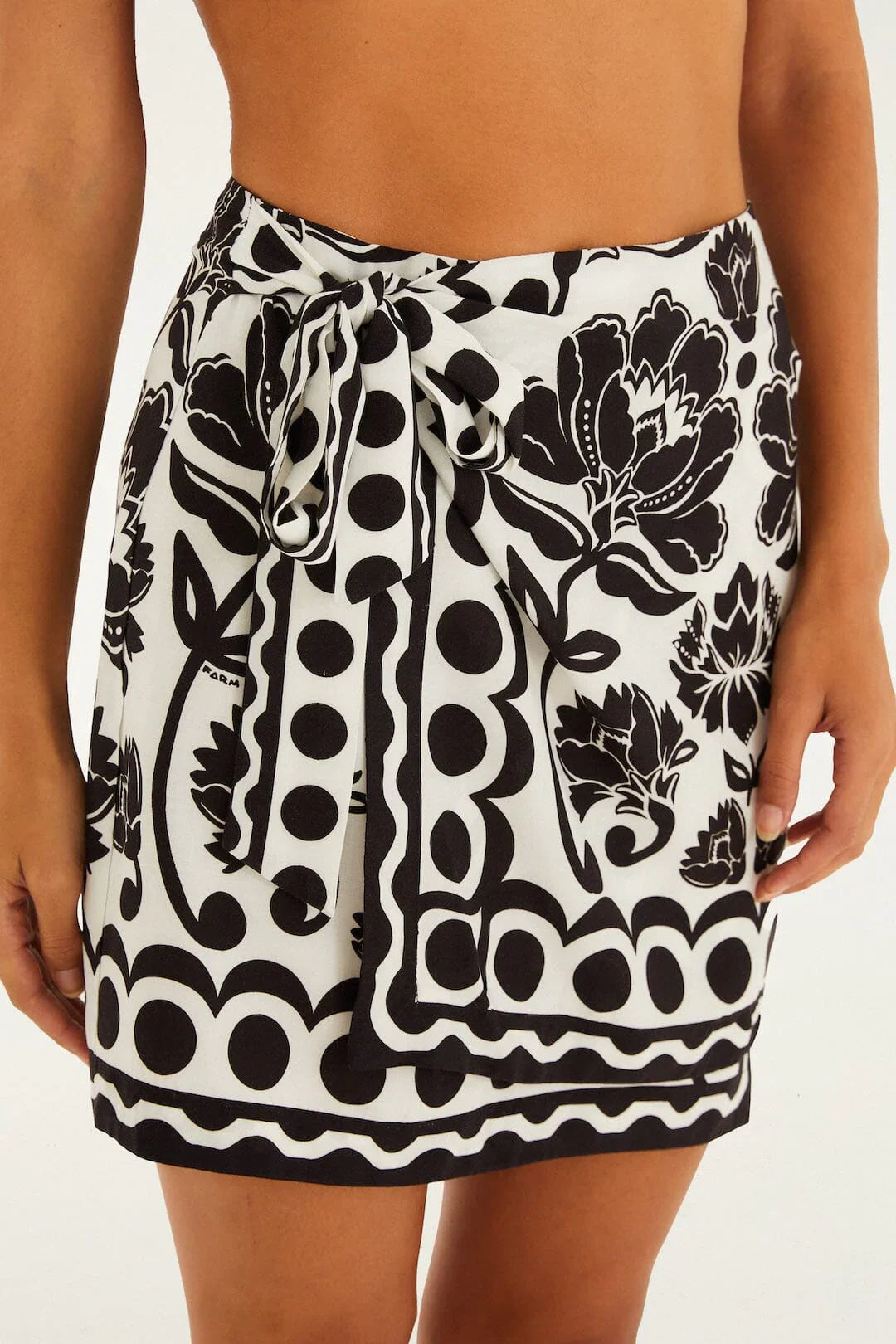 A person models the Zertz FLORAL ARABESQUE MINI SKIRT—a black and white floral wrap skirt with a waist-tied bow—shown from mid-torso to mid-thigh against a plain background.