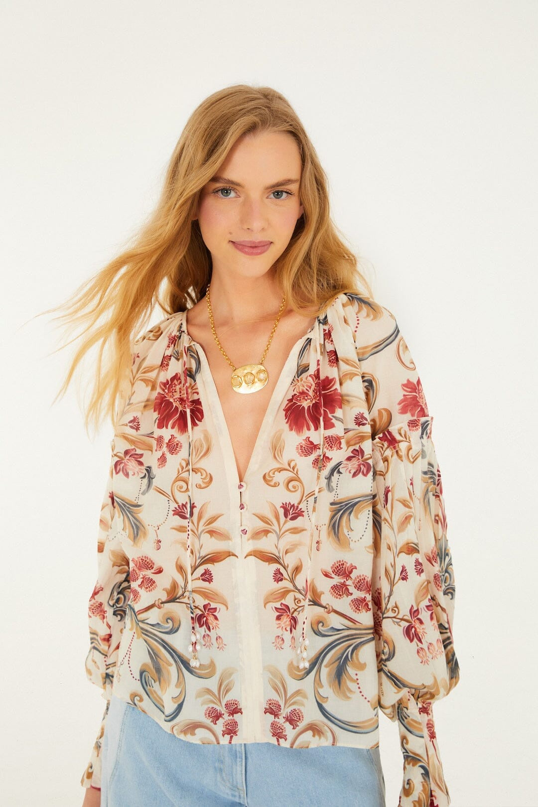 A woman with long blonde hair wears a Zertz ROYAL GARDEN SAND BLOUSE in red and beige floral print, paired with light blue jeans and a gold necklace, standing against a plain white background.