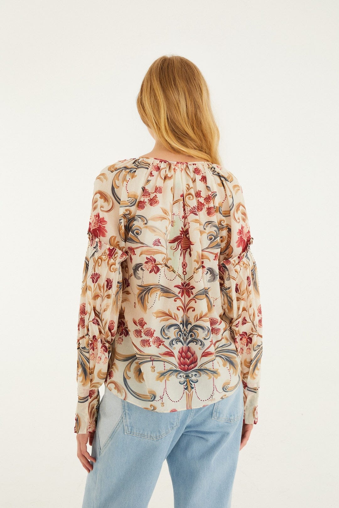 A woman with long blonde hair, seen from behind, wears a light-colored Zertz ROYAL GARDEN SAND BLOUSE featuring red, blue, and beige floral and paisley patterns, paired with light blue jeans against a white background.