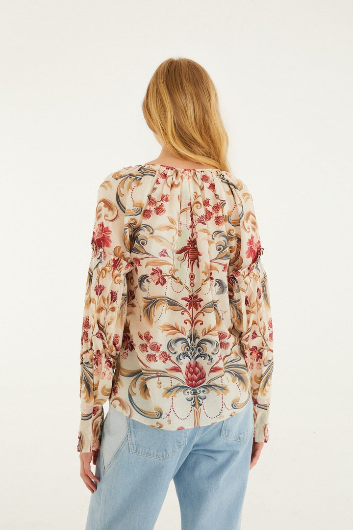 A woman with long blonde hair, seen from behind, wears a light-colored Zertz ROYAL GARDEN SAND BLOUSE featuring red, blue, and beige floral and paisley patterns, paired with light blue jeans against a white background.