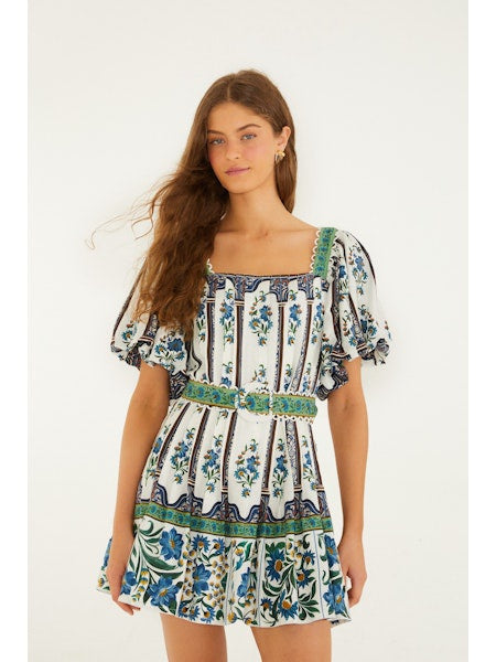 A young woman with long brown hair models the Zertz MANOELA SHORT SLEEVE BELT DRESS—a short white dress with blue and green floral prints, puffed sleeves, a square neckline, and ruffled hem—against a plain white background.