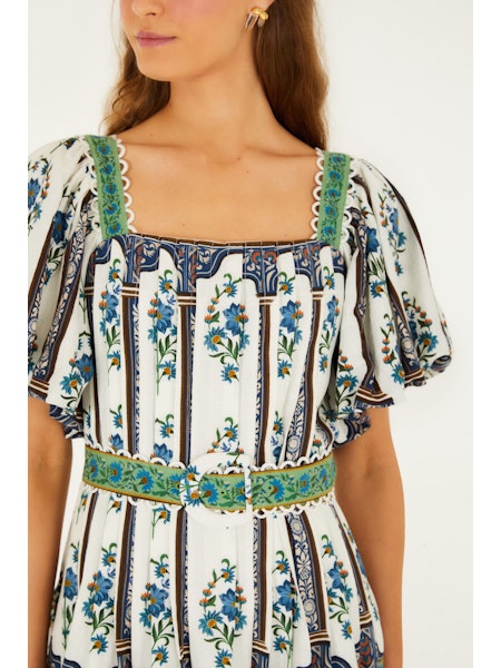 A woman models the Zertz MANOELA SHORT SLEEVE BELT DRESS featuring blue floral patterns, green lace trim, and puffed sleeves. The flattering white dress is shown from shoulders to waist against a plain background.