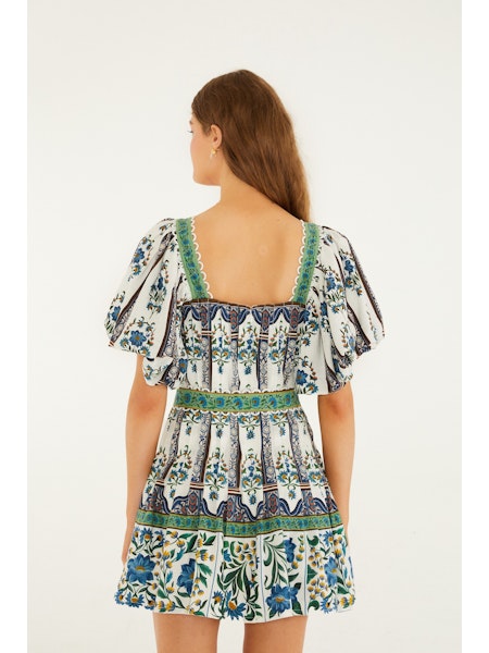 A woman with long brown hair is seen from the back wearing the Zertz MANOELA SHORT SLEEVE BELT DRESS, a white floral-patterned piece featuring green and blue accents, intricate designs, flutter sleeves, and a square neckline.