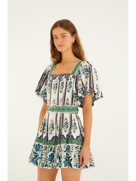 A woman models the Zertz MANOELA SHORT SLEEVE BELT DRESS, featuring a white base with blue and green florals, flutter sleeves, a square neckline, and a flared skirt—an effortlessly flattering essential for any wardrobe.