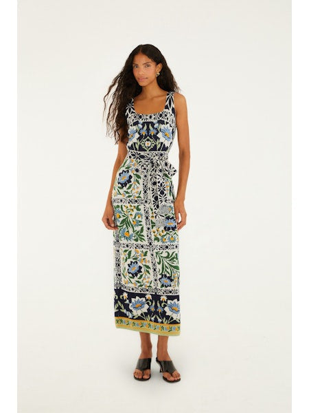 A woman with long curly hair wears the Zertz MANOELA KNIT MIDI DRESS—a sleeveless, ankle-length design with a colorful floral and geometric pattern and belted waist—paired with black sandals, standing against a white background.